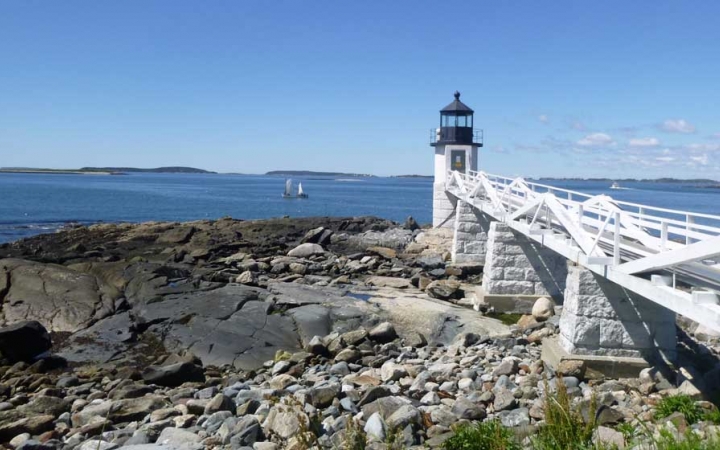 A white wooden dock stretches over a rocky beach to a lighthouse on the shore's point. Beyond the lighthouse is blue water, with a ship floating in the distance.